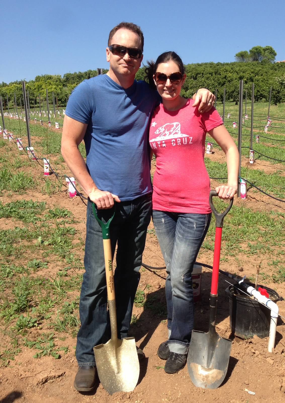 man and women both holding shovels, vineyard in the background 